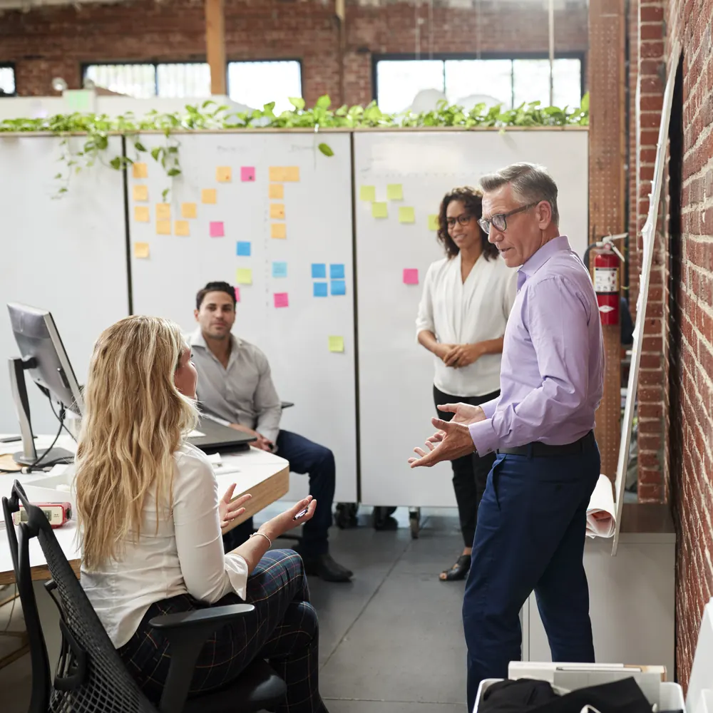 A working session at a wall of whiteboards and sticky notes: one person explains while three colleagues listen